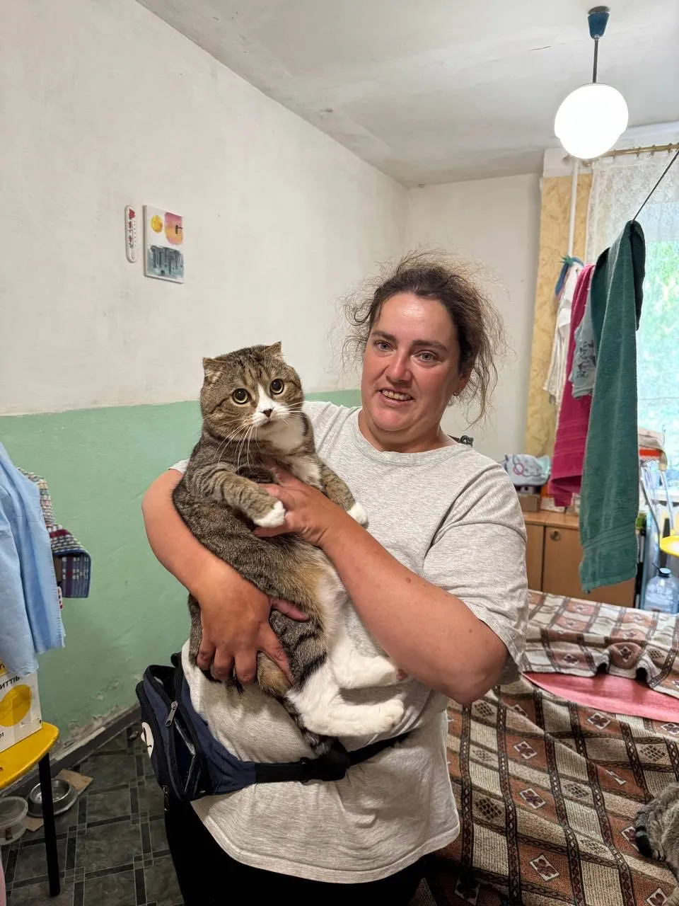 An Ukrainian woman smiles while holding a large cat with folded ears and white markings, both looking towards the camera in an indoor setting.