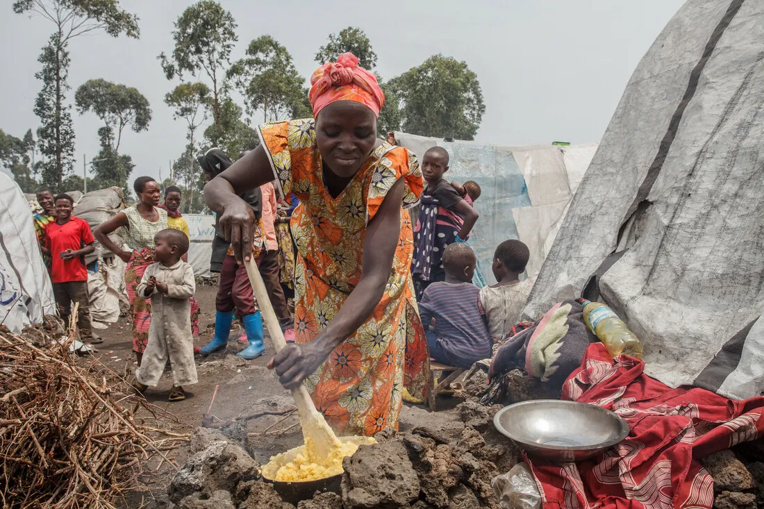 A woman preparing food outside a makeshift shelter, with children nearby and similar shelters in the background.