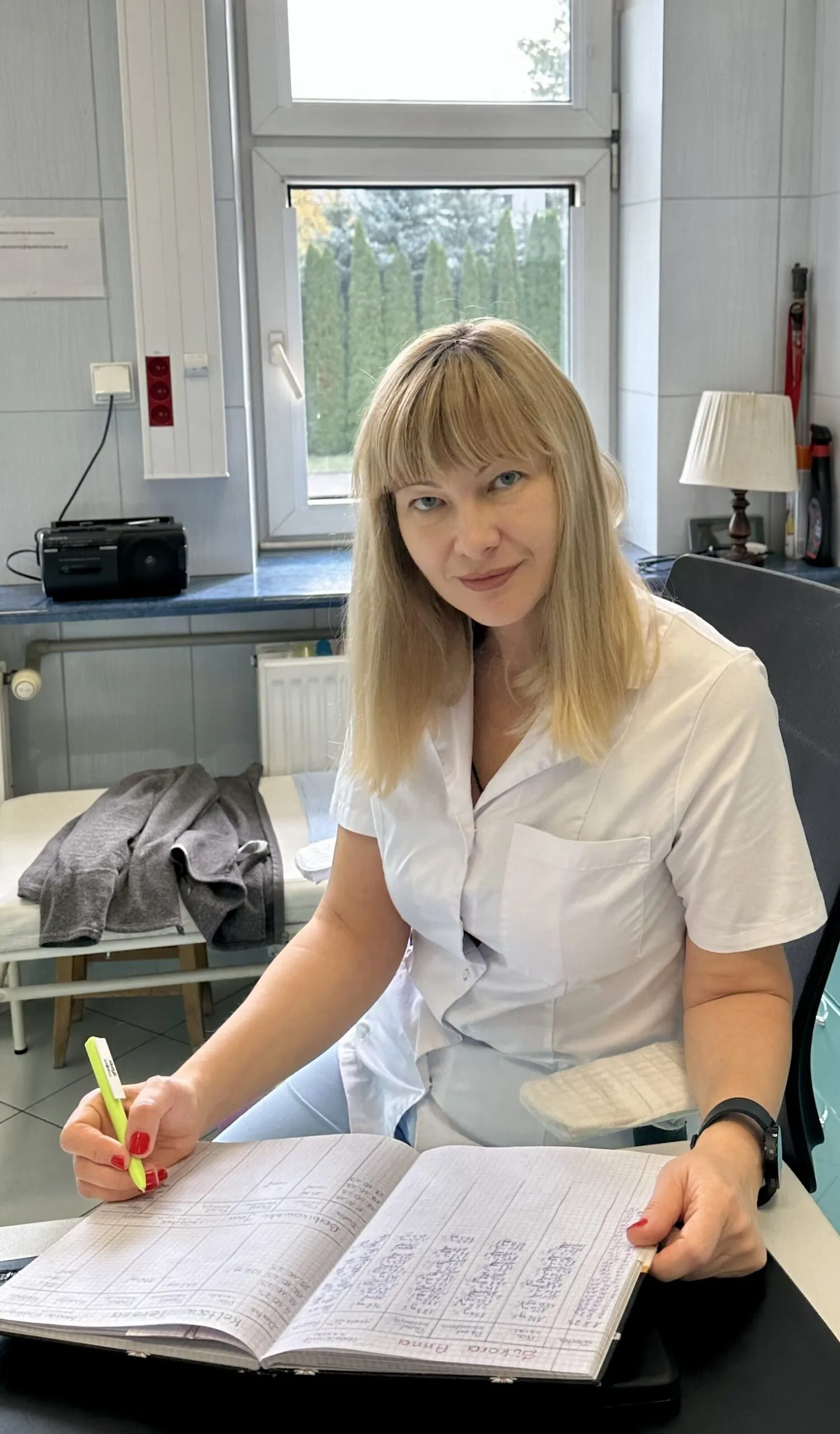 An Ukrainian woman in a white lab coat smiles at the camera while writing in a ledger in an office with medical equipment and a window showing trees.