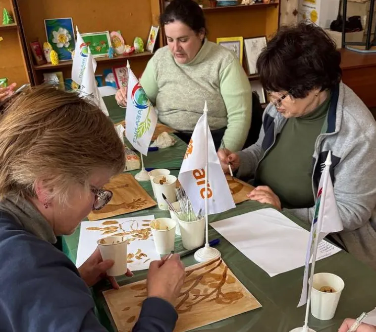 Three internally displaced Ukrainian women of different ages are intently painting with watercolors, focusing on their respective artworks. A CARE logo flag is visible in the center of the table.