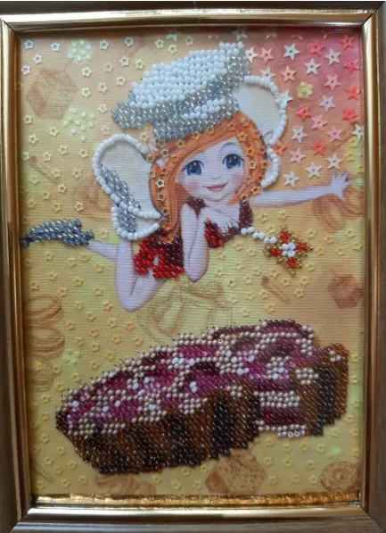 A framed beaded artwork depicts a smiling cartoon-like angel with red hair, white wings, and a chef's hat, floating above a richly textured, bead-embroidered cake or loaf. The background is a golden fabric adorned with small stars and geometric patterns.