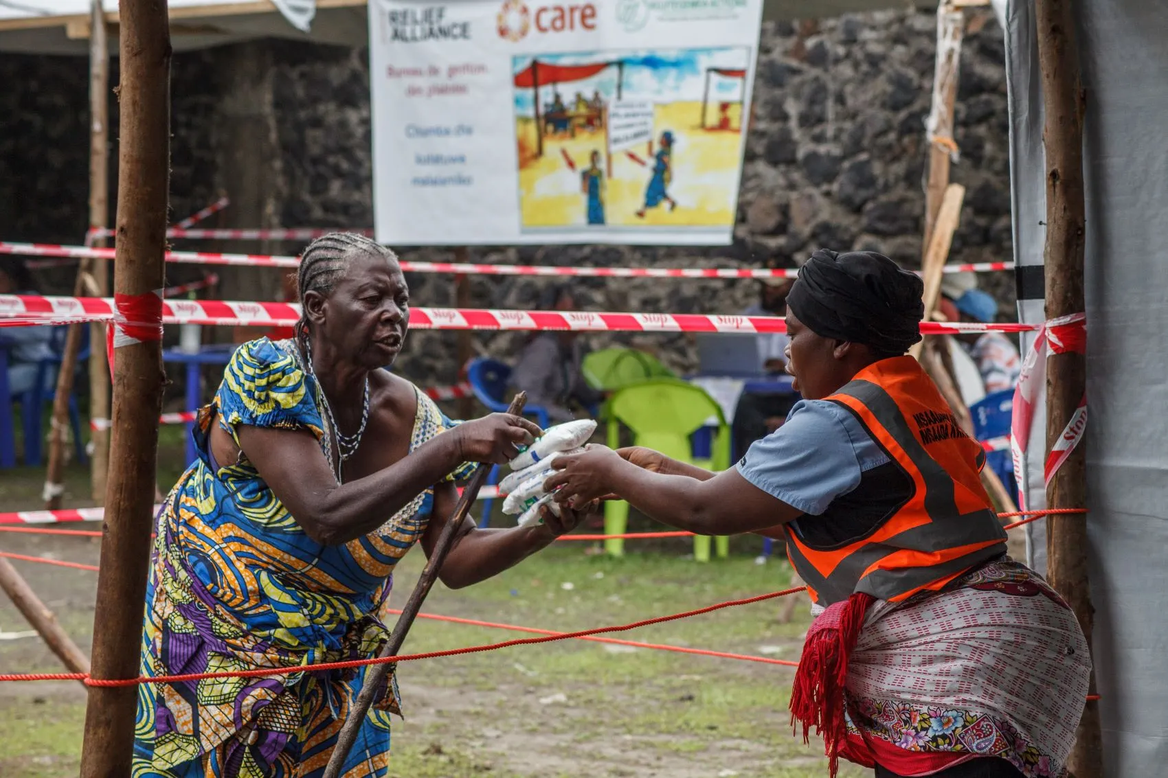 A woman receives food assistance from another woman wearing an emergency vest, likely a humanitarian worker.
