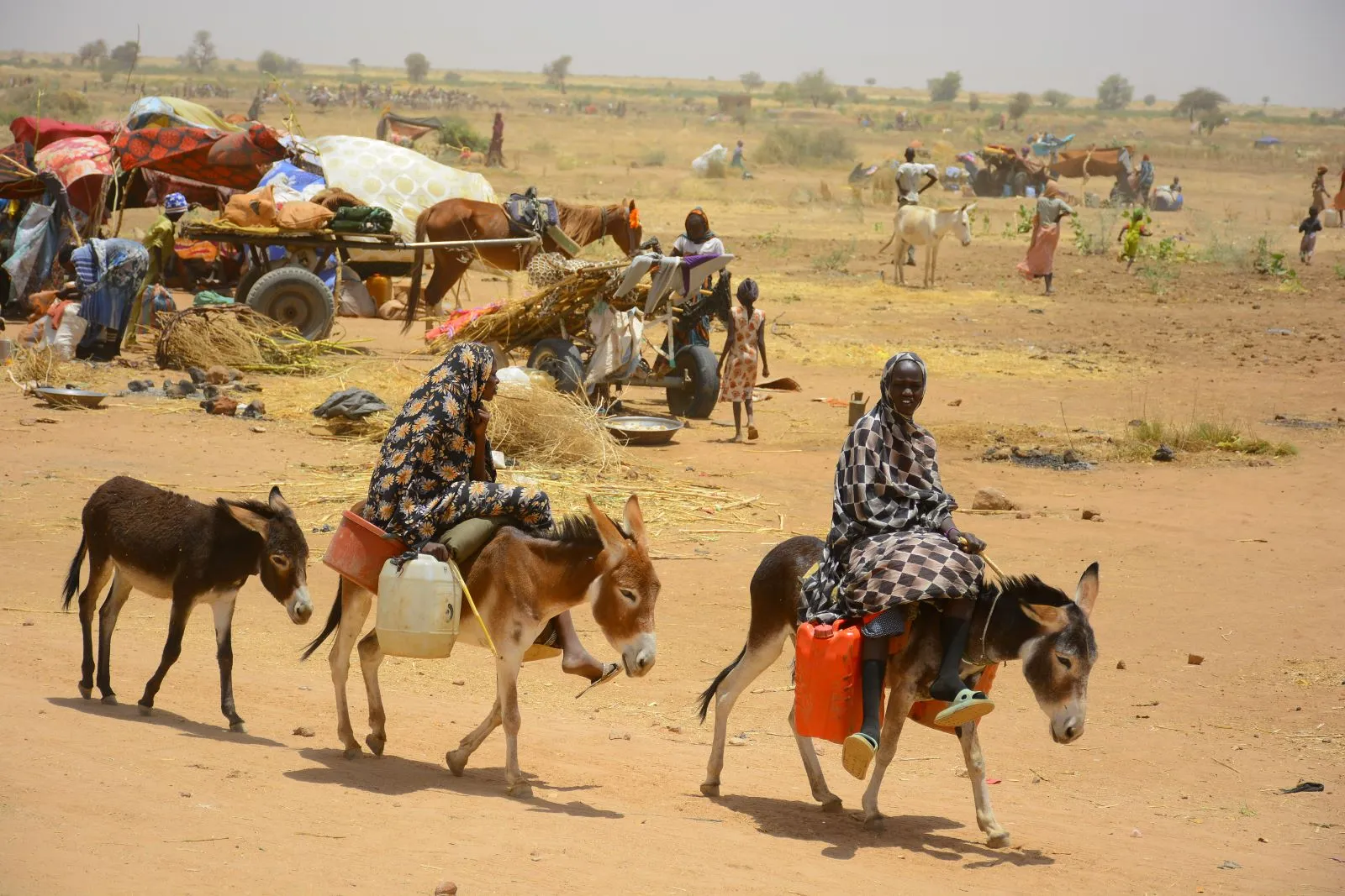 Two displaced Sudanese women ride donkeys carrying water jerrycans, with other families, carts, animals, and belongings visible in the background.