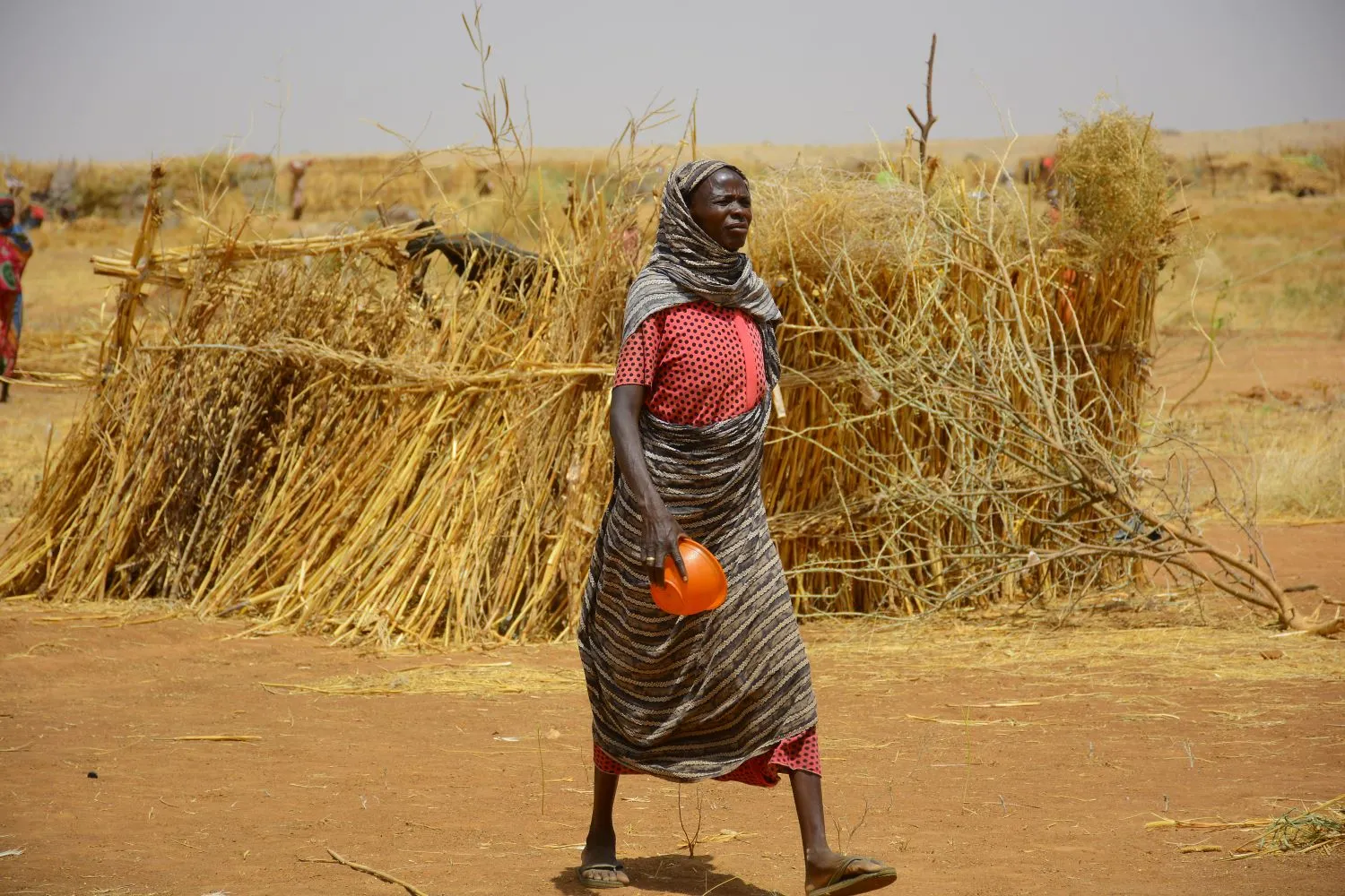 Sudanese woman walking, holding a plastic pot, with a thatched shelter visible behind her.