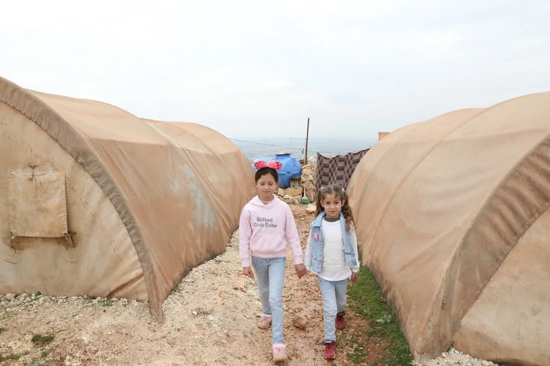 Two young girls confidently walk towards the camera between two makeshift tent shelters.