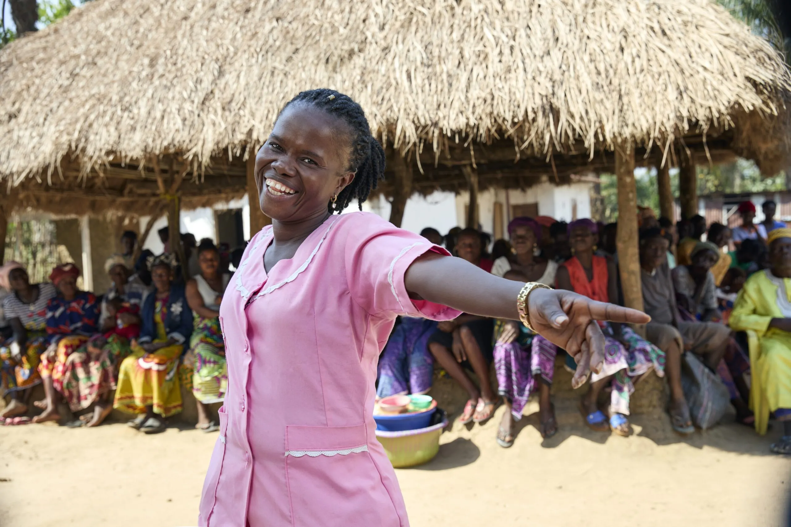 A woman wearing pink scrubs dances in the middle of a village.