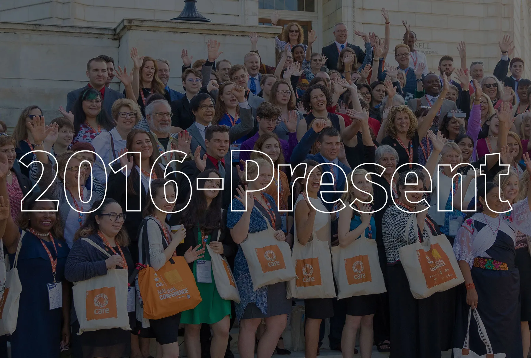 A large group of men and women, some sporting CARE canvas tote bags, raise their hands and cheer and smile.
