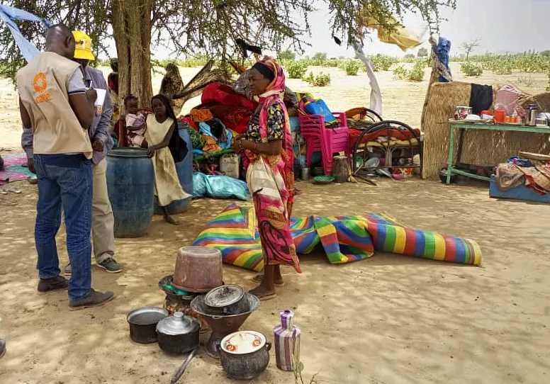CARE staff in vest writes on clipboard in outdoor setting while several people talk to him.