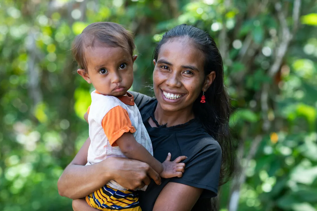 A woman smiles while holding up her young child. They are surrounded by lush plants and trees.