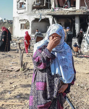 A woman wearing a headscarf covers her face with her hand. Behind her, people walk among rubble.