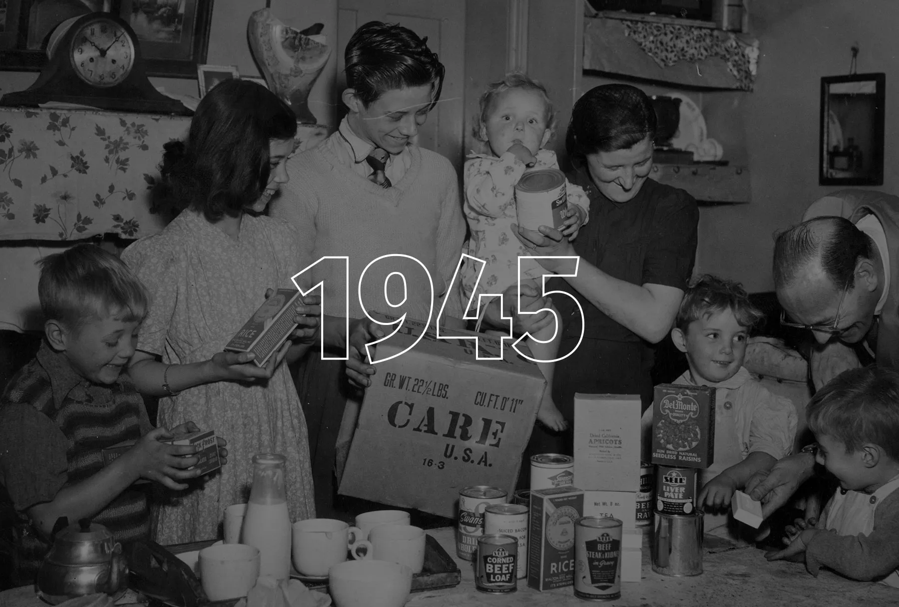 A large family convenes around a table filled with food items from a CARE Package.