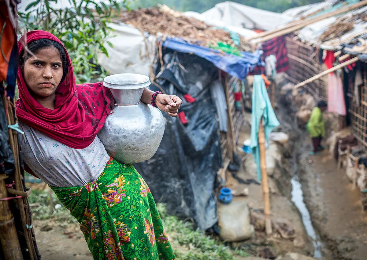 A woman wearing bright pink and bright green rests against a post. She is holding a large silver jug in her arm. Behind her are a row of tents covered in tarps.