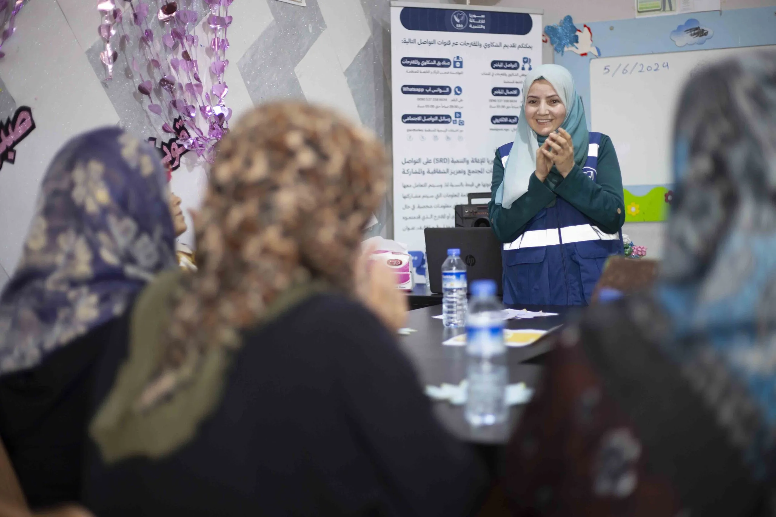 A woman in hijab and a reflective vest smiles at three other women, all in hijab, during a group session at the SRD center.