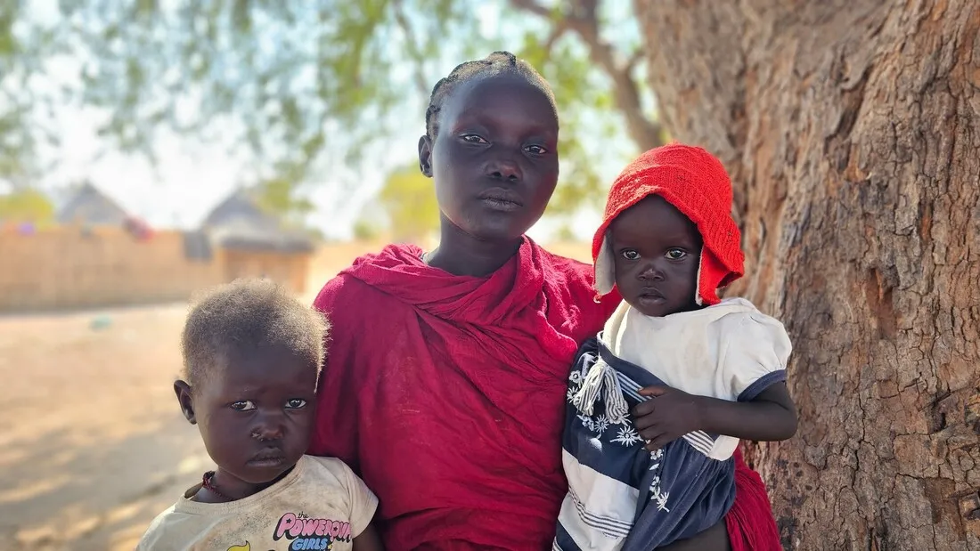 A woman holds two small children beneath the shade of a tree.